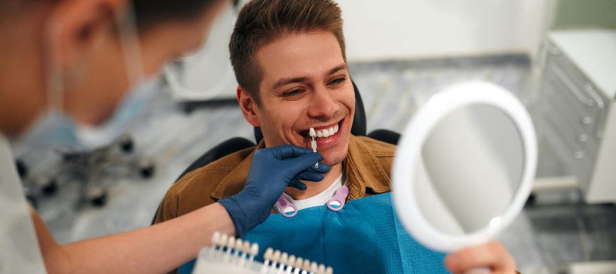 Medicine, dentistry and healthcare concept - closeup of a dentist with tooth color samples choosing a shade for a male patient's teeth in a dental clinic looking at a mirror.